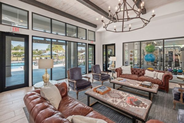 Clubhouse interior featuring modern decorations and ample lounging including two couches, four armchairs, two coffee tables, and a view into the fitness center and poolside sundeck through large clear windows